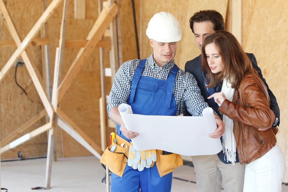 Chantier de construction résidentielle avec architecte ou contremaître en casque blanc et salopette bleue présentant des plans à un couple de clients